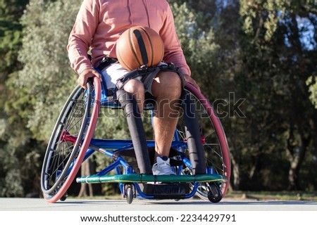Man of unrecognizable age sitting in sport wheelchair. Person with prosthetic leg having basketball on lap going for training outdoors on sunny day. Sports equipment, amputee sport concept