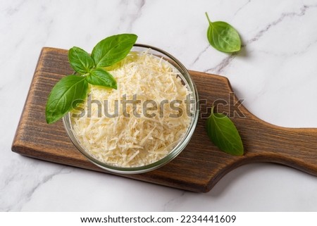 Bowl of grated grana padano cheese over marble surface. Finely shredded parmesan in a glass bowl and green basil prepared for cooking. Delicious dairy product. Italian hard cheese. Top view. Royalty-Free Stock Photo #2234441609