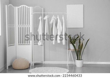 Rack with stylish sweaters, houseplants and folding screen near light wall in room