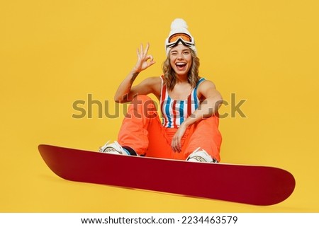 Snowboarder satisfied woman wear orange ski suit goggles mask hat swimsuit spend extreme weekend sitting show ok gesture wink isolated on plain yellow background. Winter sport hobby trip relax concept
