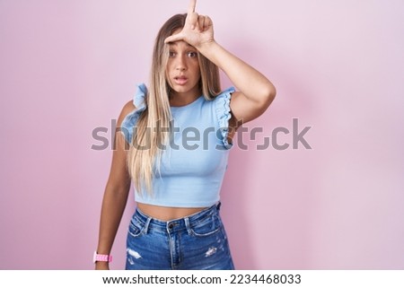 Young blonde woman standing over pink background making fun of people with fingers on forehead doing loser gesture mocking and insulting. 