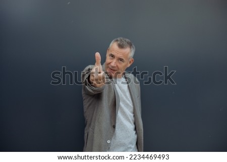 Close up serious, strong, confident grizzled man looking at camera, showing, presenting by index finger to camera some new product, service against blue wall. Place for advertisement text. Copy space