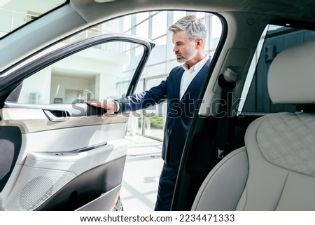 Serious thoughtful middle aged caucasian man choosing new car in dealership. Happy beard grey hair man finally gets long-awaited car, wearing formal suit. Royalty-Free Stock Photo #2234471333