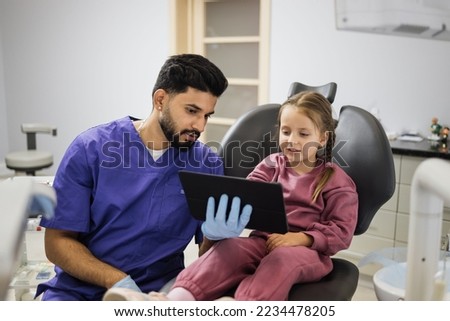 Medicine, pediatric dentistry and oral care concept. Confident male smiling professional dentist showing tablet pc computer to happy kid patient, caucasian preschool girl at modern dental clinic.