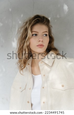 Stylish woman standing against a white wall in a rays of the sun with beautiful shadows. A girl in beige fashionable clothes leaning against the wall. Portrait of a young beautiful woman, wavy hairs  