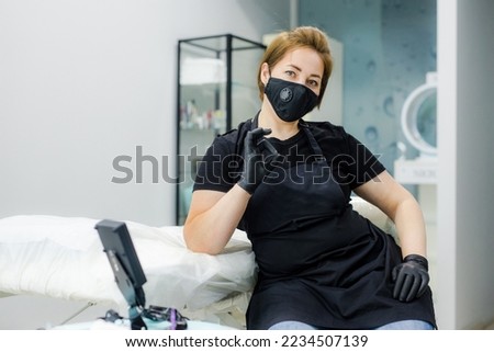 A woman in a protective mask and rubber gloves sits in a chair near a professional sofa. Beautiful girl is going to get permanent makeup in her beauty studio. The person is tattooing. Royalty-Free Stock Photo #2234507139