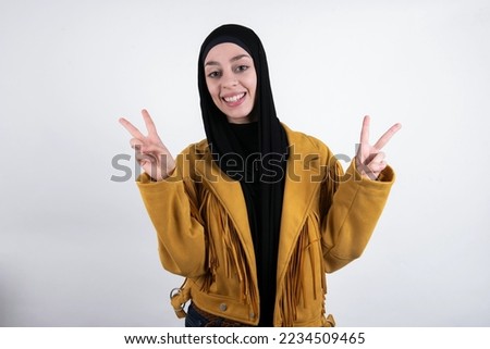 Isolated shot of cheerful young beautiful muslim woman wearing hijab and yellow jacket over white background makes peace or victory sign with both hands, feels cool.