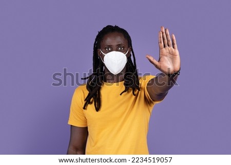 Concerned african american young man with protective mask of coronavirus disease holding hand in stop sign, warning from COVID-19, looking at camera with a worried expression, purple background