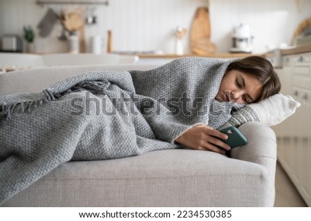 Upset sad teenage girl lying under blanket on sofa holding phone, kid spending time staring at screen, sick child using mobile phone while resting in bed at home. Gadget addiction among children Royalty-Free Stock Photo #2234530385