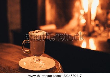 Hot chocolate in a glass on wooden table with cozy fireplace flame on the background. Winter season.