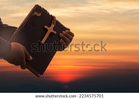 Hands holding Old Holy Bible book and Cross on sunset sky. Religion and Hope Concept.  Royalty-Free Stock Photo #2234575701