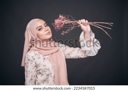 Close up model wearing floral dress with hijab, holding a dried flower isolated over dark background. Stylish Muslim female fashion lifestyle  concept.