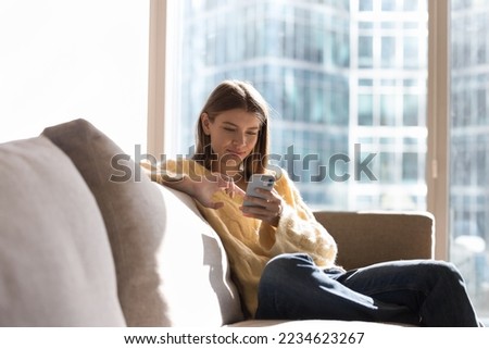 Happy thoughtful millennial gen Z girl using mobile phone at home, sitting on home couch with big window, natural day light in background, enjoying shopping, online communication