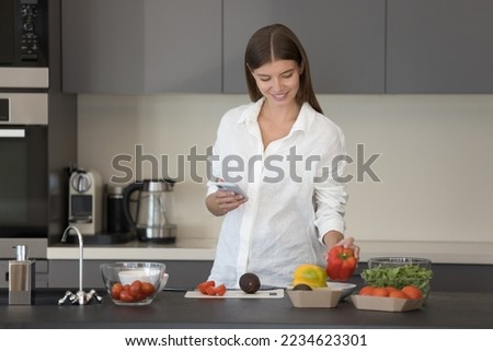 Happy pretty young Caucasian woman consulting Internet recipe, cook blog, using smartphone, preparing salad for dinner, cutting fresh vegetables on home kitchen table, smiling