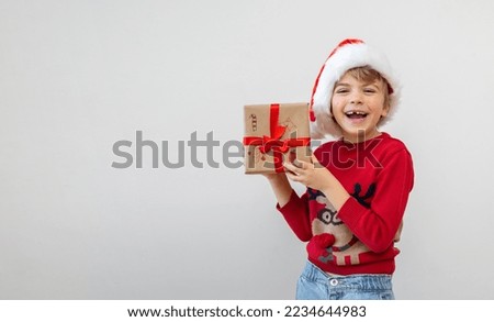 Excited funny caucasian child girl wear red Santa hat holding Christmas gift boxes, dreaming over white background. Copy space. Merry Christmas presents shopping sale.