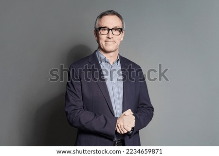 Mature confident businessman in suit, checked blue and white shirt and glasses is standing on grey studio wall background
