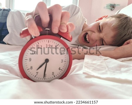 Stressed tired boy sleeping in bed with an alarm clock. Morning and time to get up laziness aggression emotions Royalty-Free Stock Photo #2234661157