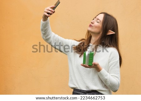 Smiling young woman with elf ears holding green gift box and taking selfie on mobile phone on the yellow wall background