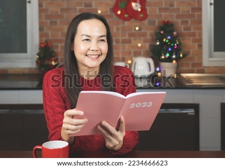 Portrait of Asian woman wearing red knitted sweater sitting  at table with red cup of coffee in the kitchen decorated with Christmas tree, holding opened 2023 planner, smiling and looking away.