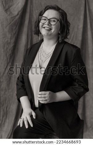A girl with a bob, black and white photo, sits on the background of the material, in a suit, jewelry and glasses, looks at the camera, portrait photo, happy