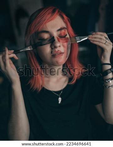 Young redhead woman artist applies red and black watercolor paint to her face with brush