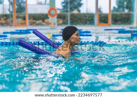 Side view indoor portrait focused caucasian senior woman in a black head cap using violet pool noodle during swimming. Equipment concept. High quality photo Royalty-Free Stock Photo #2234697577