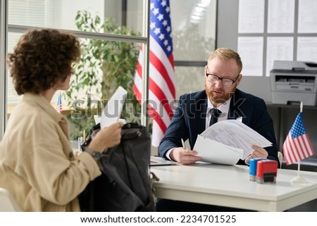 Portrait of male worker revising documents of young woman applying for visa in US immigration office Royalty-Free Stock Photo #2234701525