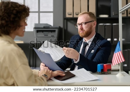 Portrait of bearded male consultant working in US embassy office and holding application form while talking to woman Royalty-Free Stock Photo #2234701527