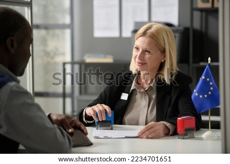 Portrait of smiling mature woman consulting person in EU immigration office or government agency and stamping forms Royalty-Free Stock Photo #2234701651