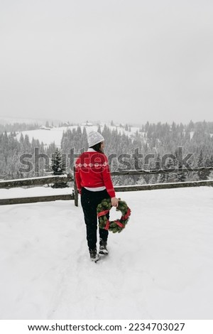 Portrait of a beautiful young woman in a red Christmas sweater and a white hat holds a New Year's holiday wreath outdoor in the winter in the snowy mountains. Vacation trip. Beautiful winter landscape