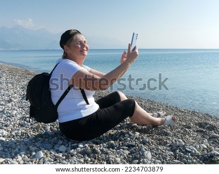 Senior adventureisageless woman with backpack taking a selfie on a mobile phone on the sea beach. Summer active tourism for pensioner. Internet for holiday concept