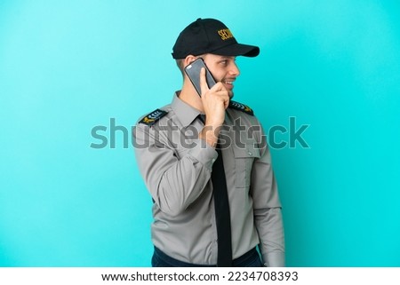 Young security man isolated on blue background keeping a conversation with the mobile phone with someone