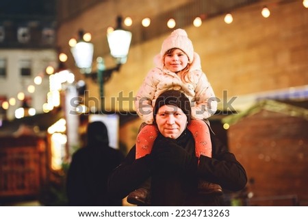 Little preschool girl sitting on shoulder of father on Christmas market in Germany. Happy toddler child and man observing traditional decorated pyramid. Happy family, bonding, love. Family xmas time