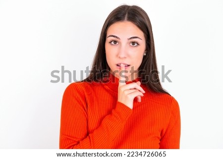 Surprised Young caucasian woman wearing red sweater over white background makes silence gesture, keeps finger over lips and looks mysterious at camera