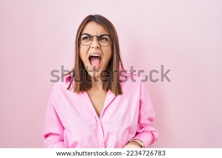 Young hispanic woman wearing glasses standing over pink background angry and mad screaming frustrated and furious, shouting with anger. rage and aggressive concept. 