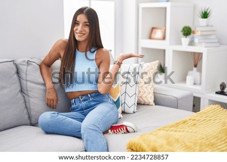 Young brunette woman sitting on the sofa at home smiling cheerful presenting and pointing with palm of hand looking at the camera. 