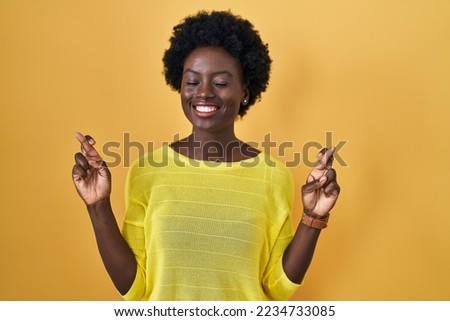 African young woman standing over yellow studio gesturing finger crossed smiling with hope and eyes closed. luck and superstitious concept. 