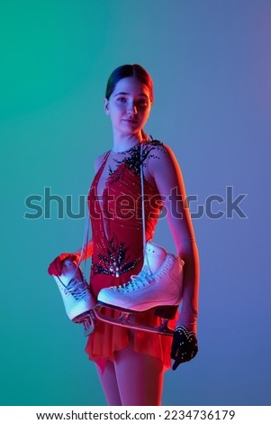 After workout. Young teen girl in stage costume dress standing with skates isolated over gradient green-blue background in neon light. Concept of skills, sport, hobbies, winter sports