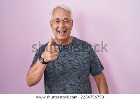 Middle age man with grey hair standing over pink background doing happy thumbs up gesture with hand. approving expression looking at the camera showing success. 