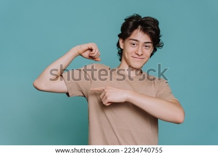 Handsome caucasian schoolboy in beige t-shirt bending arm demonstrates biceps pointing at it by index finger of another hand stands against turquoise backdrop. Cheerful Italian teen shows his strength Royalty-Free Stock Photo #2234740755