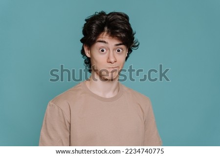 Close up portrait of surpised schoolboy in beige t-shirt standing against turquoise studio backdrop with wide opened eyes. Mockup, perplexed guy looks at camera in wondering. Promo, sale, discount. Royalty-Free Stock Photo #2234740775