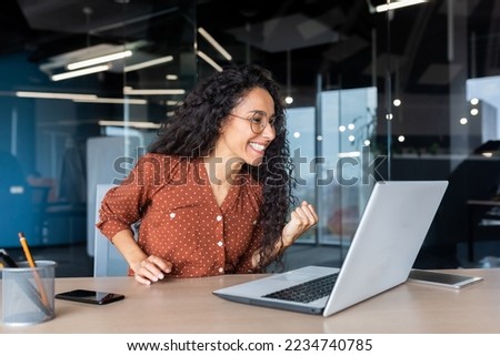Hispanic business woman celebrating victory success, employee with curly hair inside office reading good news, using laptop at work inside office holding hand up and happy triumph gesture. Royalty-Free Stock Photo #2234740785