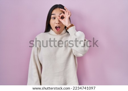 Young south asian woman standing over pink background doing ok gesture shocked with surprised face, eye looking through fingers. unbelieving expression. 