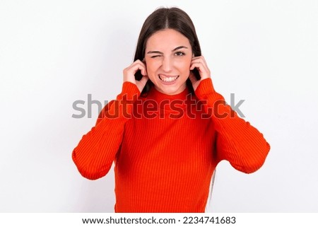 Happy Young caucasian woman wearing red sweater over white background ignores loud music and plugs ears with fingers asks to turn off sound