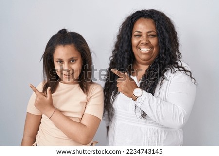 Mother and young daughter standing over white background cheerful with a smile on face pointing with hand and finger up to the side with happy and natural expression 