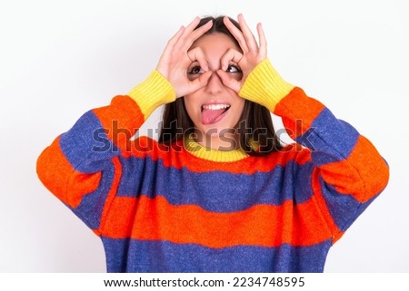 Young caucasian woman wearing colorful sweater over white background doing ok gesture like binoculars sticking tongue out, eyes looking through fingers. Crazy expression.