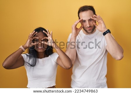 Interracial couple standing over yellow background trying to open eyes with fingers, sleepy and tired for morning fatigue 