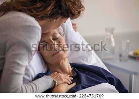 Close up of lovely daughter visiting and kissing sick mother on forehead while resting in hospital bed. Senior patient laying on gurney with eyes closed while daughter assuring a speedy recovery.  Royalty-Free Stock Photo #2234751769