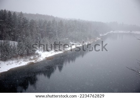 Falling snow flakes above the lake. Winter landscape with forest on the lake shore during the snowfall, selective focus. High quality photo