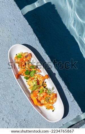 Tempura shrimp with vegetable tabouleh and spices lie in an oval ceramic plate. The plate stands on a concrete stand near the pool. Nearby is a lavender bush.